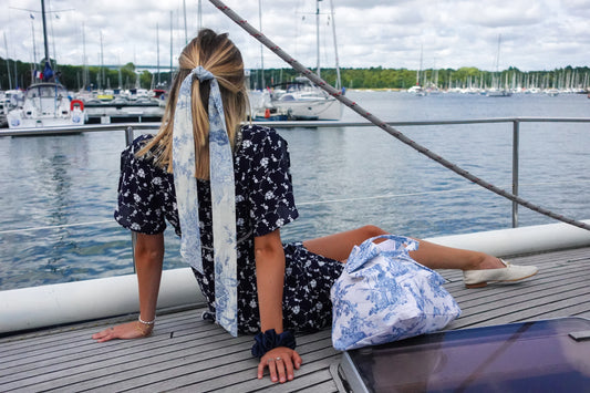 BAZAR Atelier Sablon: Foulards Jouy de Fruits en toile de Jouy bleu porté en bandeau dans les cheveux d'une jeune fille blonde. Elle est assise sur sur un voilier à quai dans un port breton. Elle porte un chouchou Soyeux bleu marine au poignet, et juste à coté d'elle se trouve un cabas bleu Jouy de Fruits.