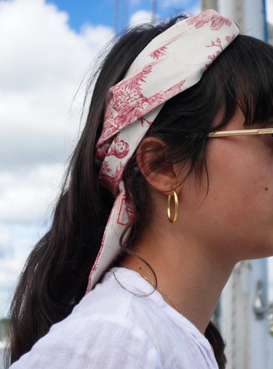 BAZAR Atelier Sablon: Foulards Jouy de Fruits en toile de Jouy rouges porté en bandeau dans les cheveux d'une jeune fille brune. Elle se trouve sur un voilier. 